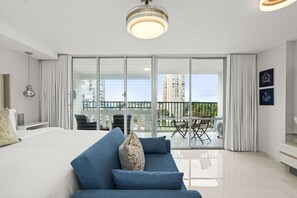 Una habitación de hotel moderna con una cama grande, un sofá azul, un balcón con mesa y sillas, y vistas a la ciudad.