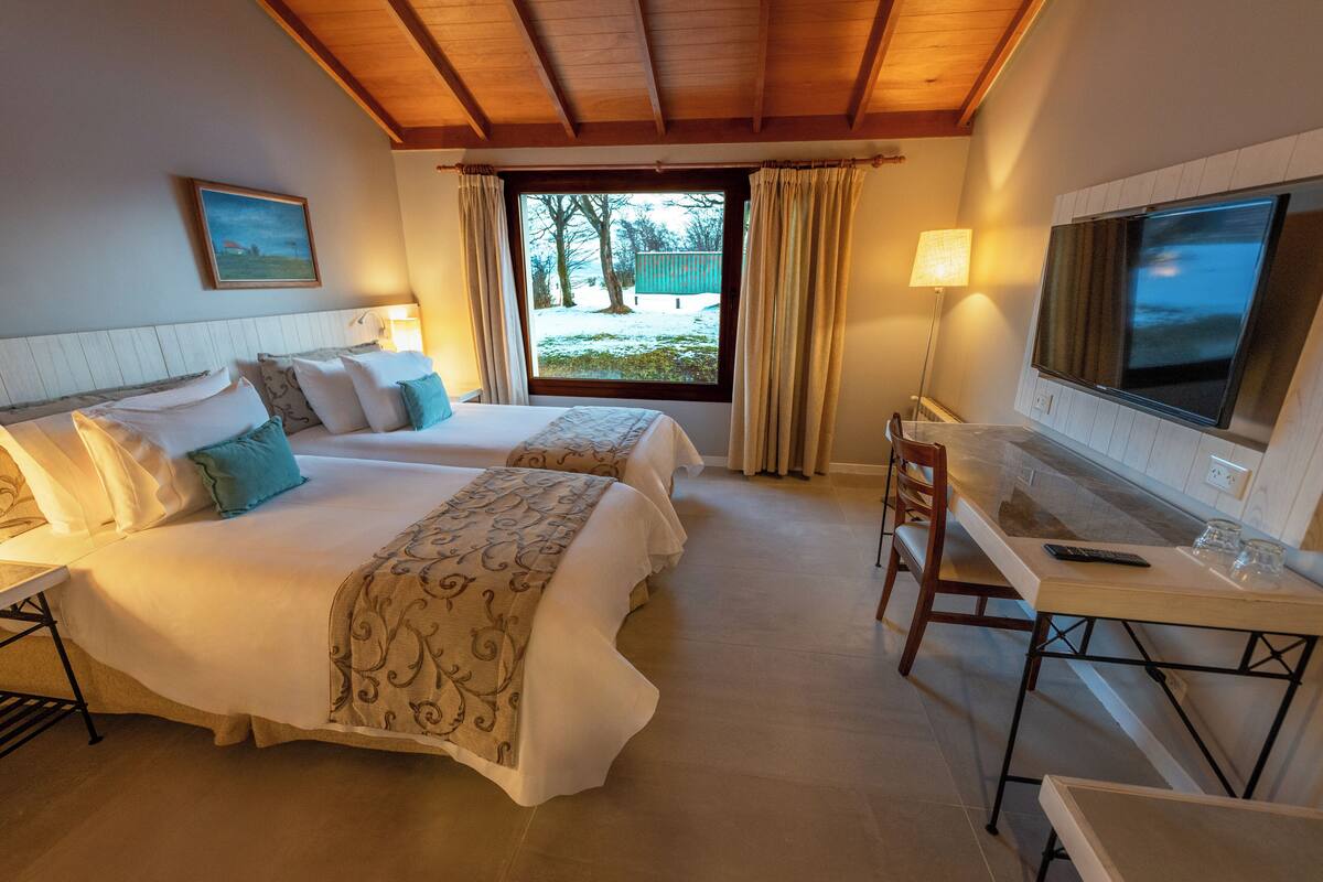 A hotel room with two beds, a desk, a TV, and a window with a view of a snowy landscape.