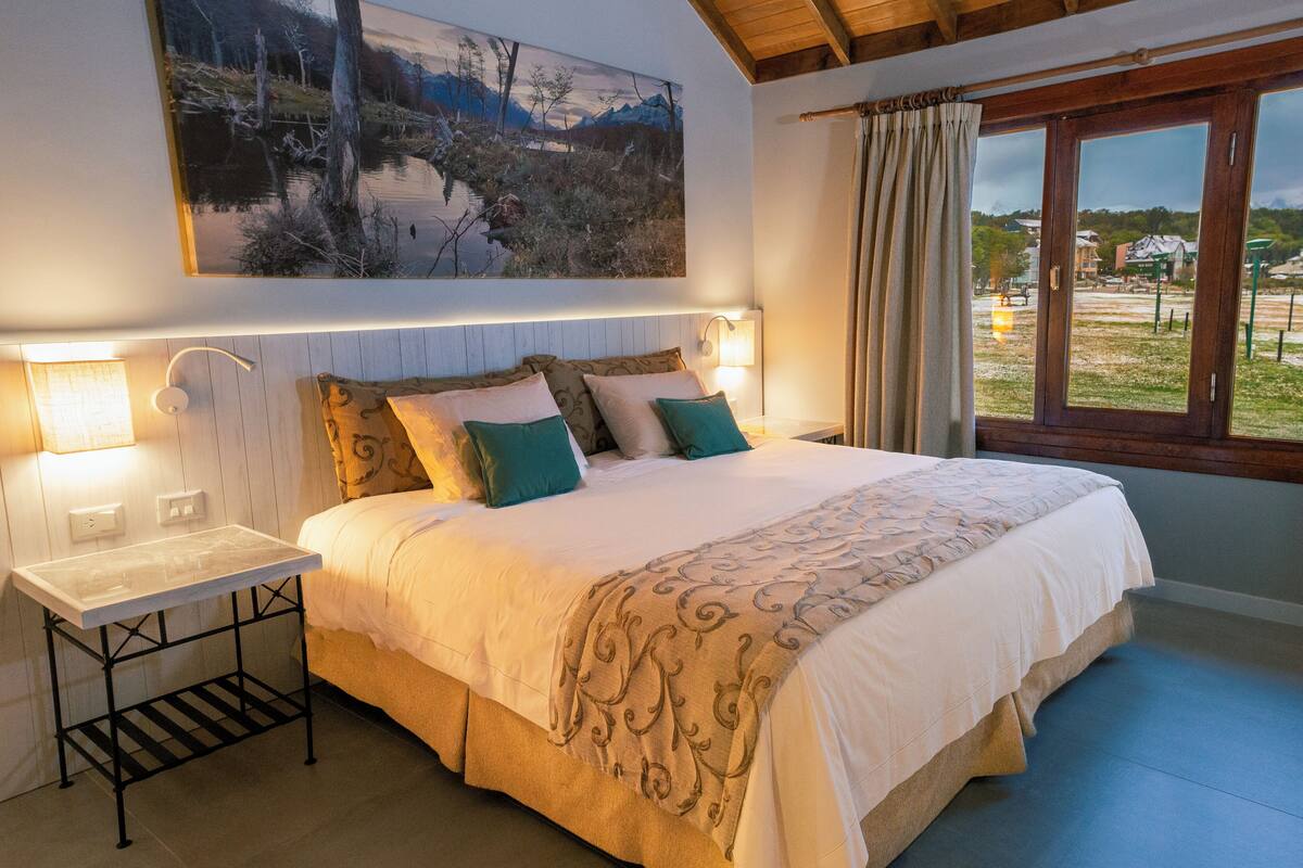 A bedroom with a bed, bedside lamps, a painting, and a view through a window.