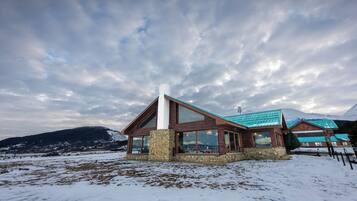 A lodge with a stone foundation, large windows, and a green roof, surrounded by snow and mountains.
