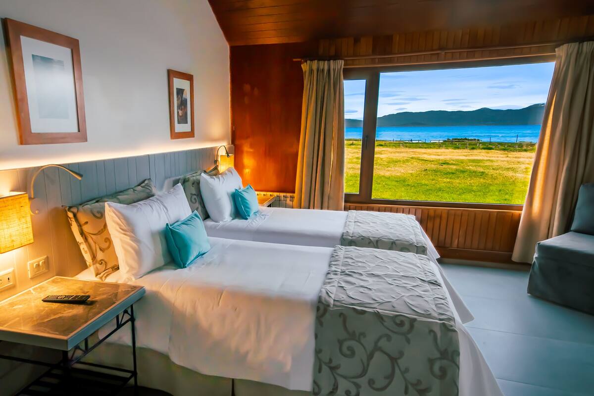 A hotel room with two beds, a desk, a lamp, and a view of the sea.