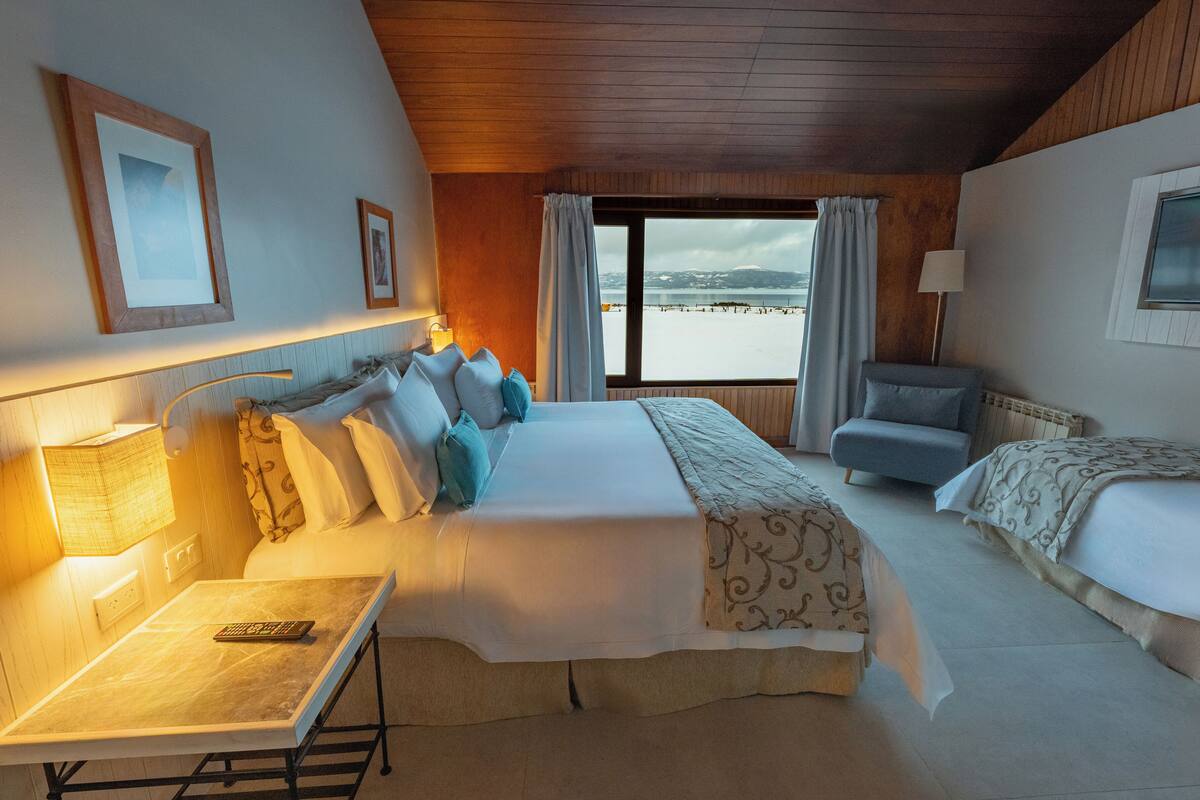 A hotel room with a large bed, a smaller bed, a desk with a lamp, a chair, and a view of a body of water through a window.