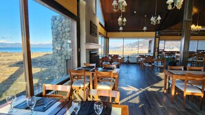 Un restaurante con mesas y sillas de madera, ventanas amplias que ofrecen vistas al paisaje y lámparas colgantes.