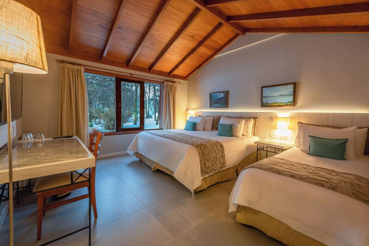 A room with two beds, a table, and a chair. There is a window with curtains, a lamp, and a painting on the wall.