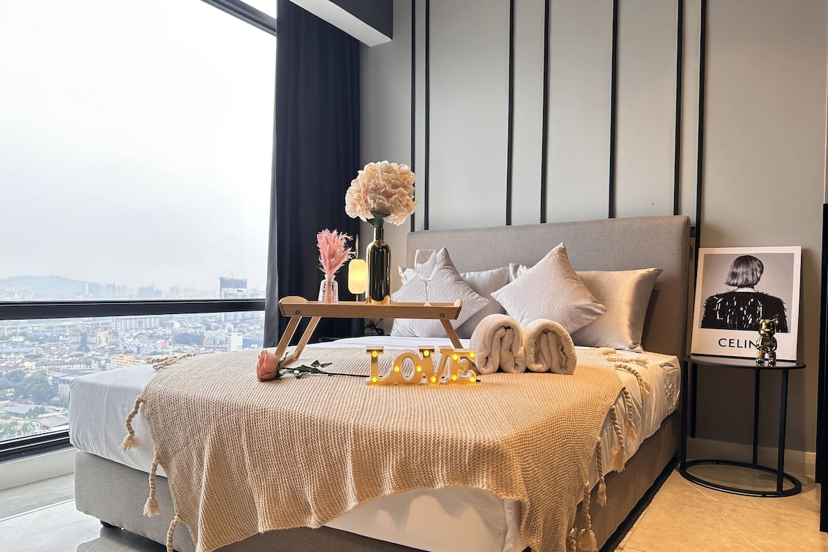 A modern bedroom with a city view, a bedside table with a vase of flowers, a framed photo, and a 'LOVE' sign.