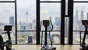 Een fitnessruimte met drie hometrainers en een loopband, grote ramen met uitzicht op een stadslandschap, waaronder de Petronas Towers.