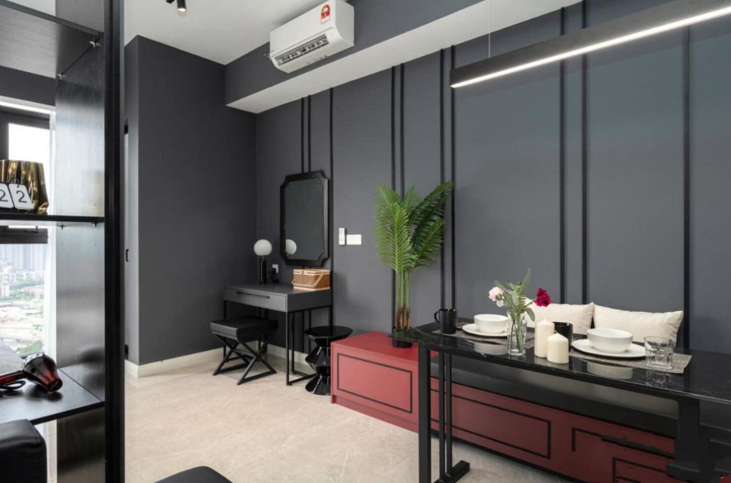Una habitación moderna con pared gris, escritorio negro, mueble rojo, silla negra, espejo, planta en maceta, sofá blanco y una mesa con un florero y velas.