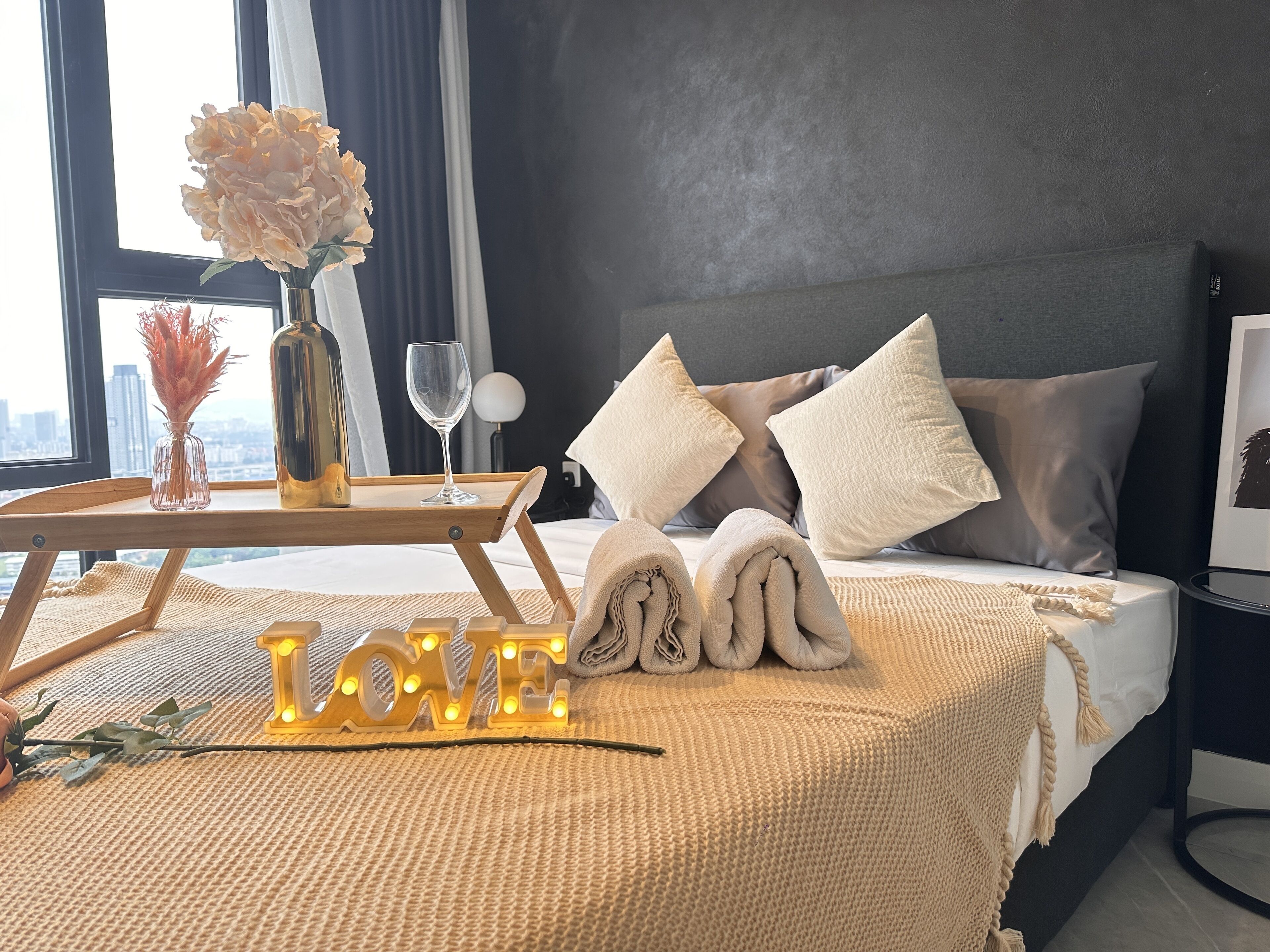 A neatly made bed with rolled towels, a tray with a vase and a wine glass, and a 'LOVE' sign on the bed.