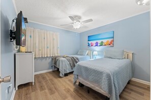 Un dormitorio con dos camas, un ventilador de techo, un cuadro con sillas de playa y un televisor sobre un soporte.