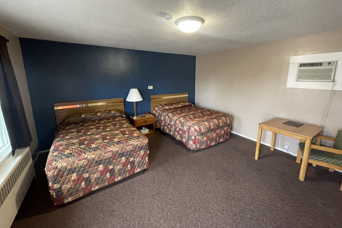 A hotel room with two beds, a small table, a lamp, and a wall-mounted air conditioner.