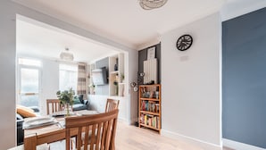 A modern living room with a dining area, featuring a wooden table and chairs, a bookshelf, and a large window with blinds.