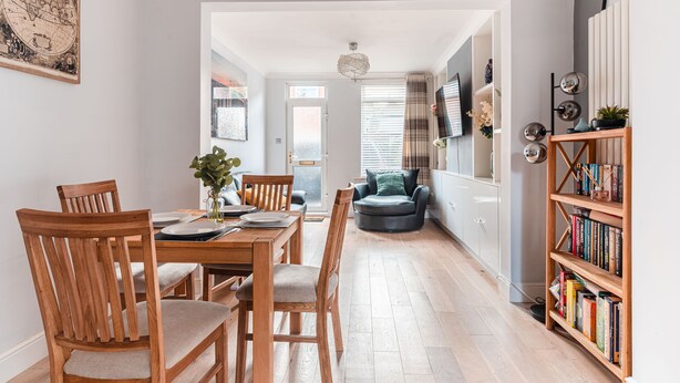 A dining area with wooden chairs and a table, a bookshelf, and a television on the wall.