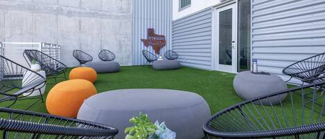Un espace extérieur aménagé avec des chaises à armature métallique noire et des poufs gris et orange posés sur un gazon synthétique, avec un mur en métal et une enseigne en arrière-plan.