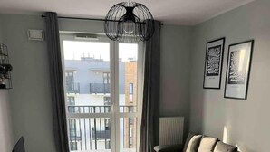 A modern living room with a sofa, a small table, and a TV. There is a large window with a view of buildings outside. - 1-bed Top Floor Apartment in Warszawa (Warsaw)