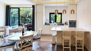 A modern kitchen with a central island, a dining area with a round table and chairs, and a view of greenery outside. - Dizengoff Brique (Tel Aviv)