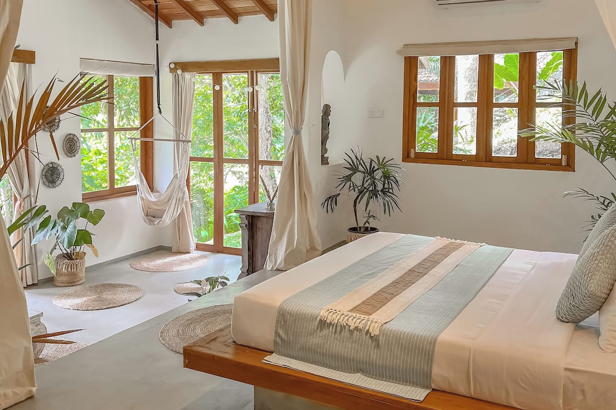 A spacious bedroom with a large bed, a ceiling fan, and a view of lush greenery outside.