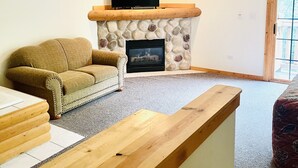 A living room with a stone fireplace, a flat-screen TV, a wooden coffee table, and a ceiling fan.