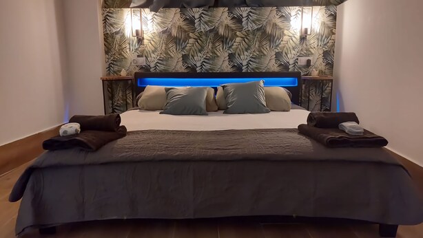 Uma cama grande com colcha cinza escura e várias almofadas, com dois mesinhas de cabeceira com abajures de cada lado.