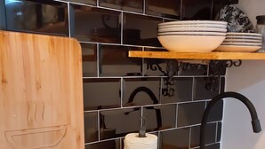 Uma cozinha com um fogão por indução preto, uma tábua de corte de madeira, um suporte para toalhas de papel, uma torneira preta e um revestimento de azulejos.