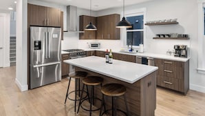 Eine moderne Küche mit Edelstahlkühlschrank, Holzschränken, einer zentralen Kochinsel mit Hockern, Pendelleuchten und einer Mikrowelle.