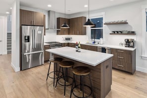 A modern kitchen with a stainless steel refrigerator, wooden cabinets, a central island with stools, pendant lighting, and a microwave.