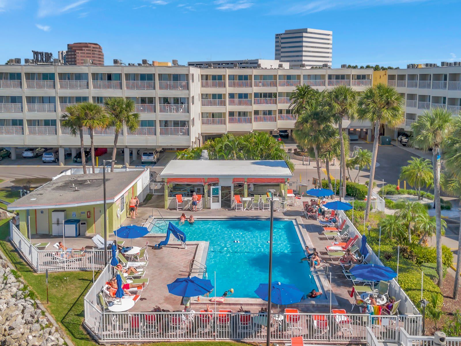 A hotel with a swimming pool surrounded by sun loungers and umbrellas, a covered area with chairs, and a parking lot with cars.