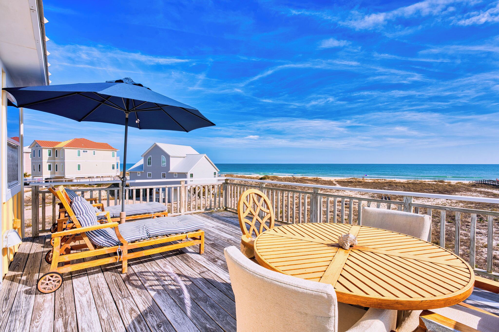 A wooden deck with lounge chairs and a table, overlooking a beach and ocean.