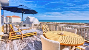 Une terrasse en bois avec des chaises longues et une table, offrant une vue sur une plage et l’océan.