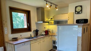 A compact kitchen with a dishwasher, a microwave, and a refrigerator. There is a window with a view of trees, a clock on the wall, and a shelf with kitchen items. - Charming Bright Spacious Village Walk to lift Hike (Praz Sur Arly)