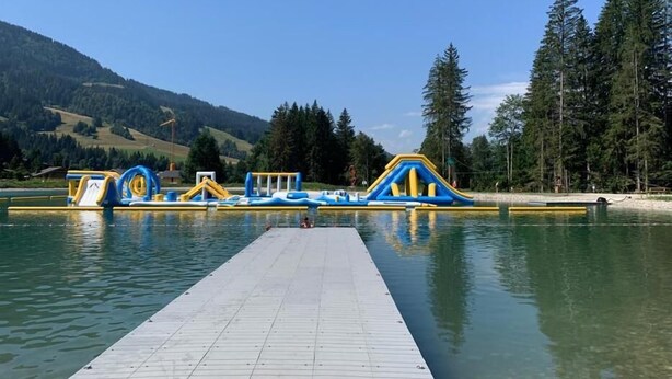 A wooden dock extending into a calm lake with inflatable play structures in blue and yellow. - Charming Bright Spacious Village Walk to lift Hike (Praz Sur Arly)