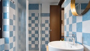 A bathroom with a checkered tile wall, a glass shower enclosure, a wooden vanity with a white sink, and a mirror with a gold frame. - Cozy, bright, comfortable penthouse, 2 min from metro Dergano.  (Milano)
