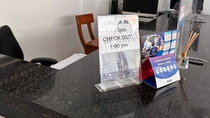 Mostrador de recepción de hotel con un cartel que indica la hora de registro a las 3:30 pm y la hora de salida a la 1:00 pm. Se ve una computadora y una silla al fondo.