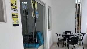 Un espacio interior limpio y bien iluminado con una puerta de vidrio que da a una zona de estar con cubiertas azules. Hay varias señales en la pared, incluyendo una que dice "NO PASE SIN CONTROL DE TEMPERATURA" y otra que dice "ZONA DE DESINFECCIÓN". Se ve una pequeña mesa con sillas en primer plano.
