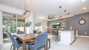 A modern kitchen with a central island, a dining area with a wooden table and blue chairs, and a large window providing natural light. - Amazing Disney resort style & FREE Waterpark 2481 (Kissimmee)