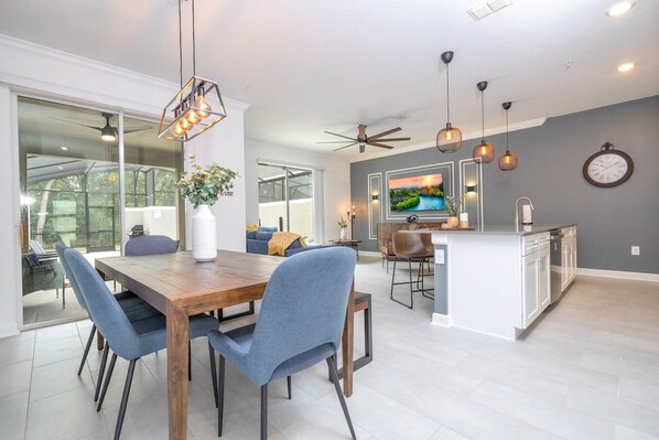 A modern kitchen with a central island, a dining area with a wooden table and blue chairs, and a large window providing natural light.