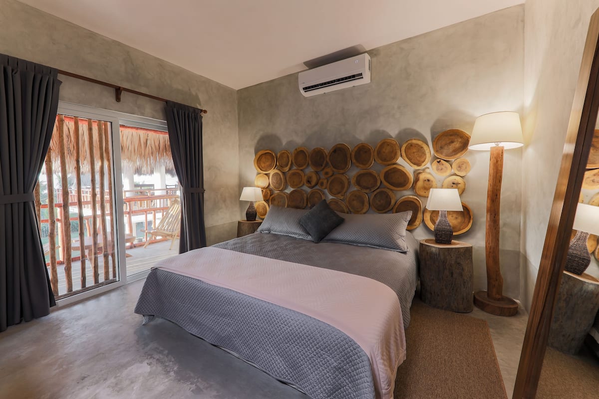 A bedroom with a large bed, a wall-mounted wooden headboard, two bedside lamps, and a view of a wooden deck through a sliding glass door.