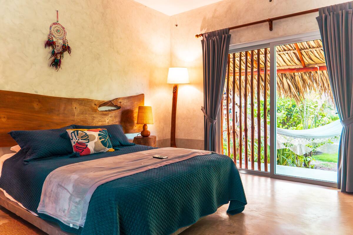 A bedroom with a large bed, a nightstand, a lamp, a dreamcatcher, and a view of a garden through a sliding door.