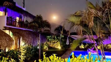 Vista nocturna de un resort con piscina, palmeras y una luna en el cielo.