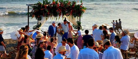A beach wedding ceremony with guests seated on the sand facing a decorated arch.