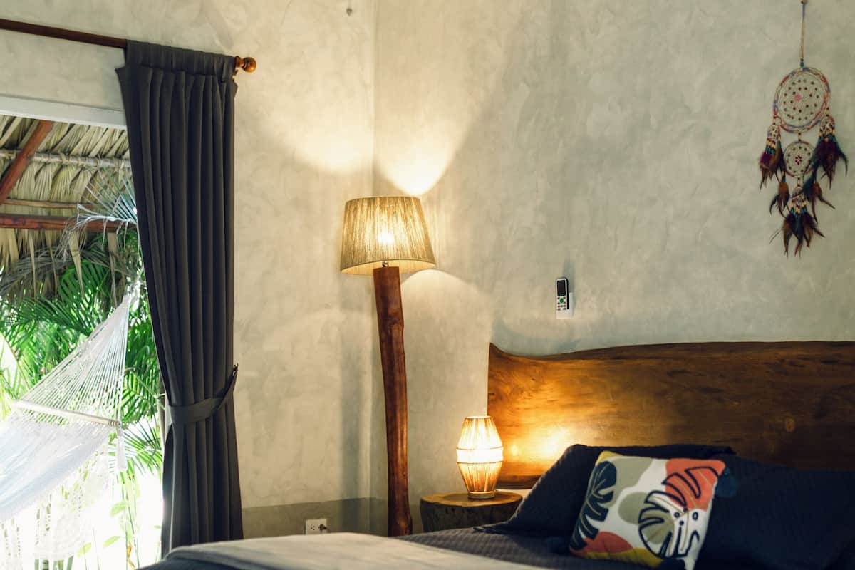 A bedroom with a bed, a nightstand with a lamp, a window with curtains, and a dreamcatcher on the wall.