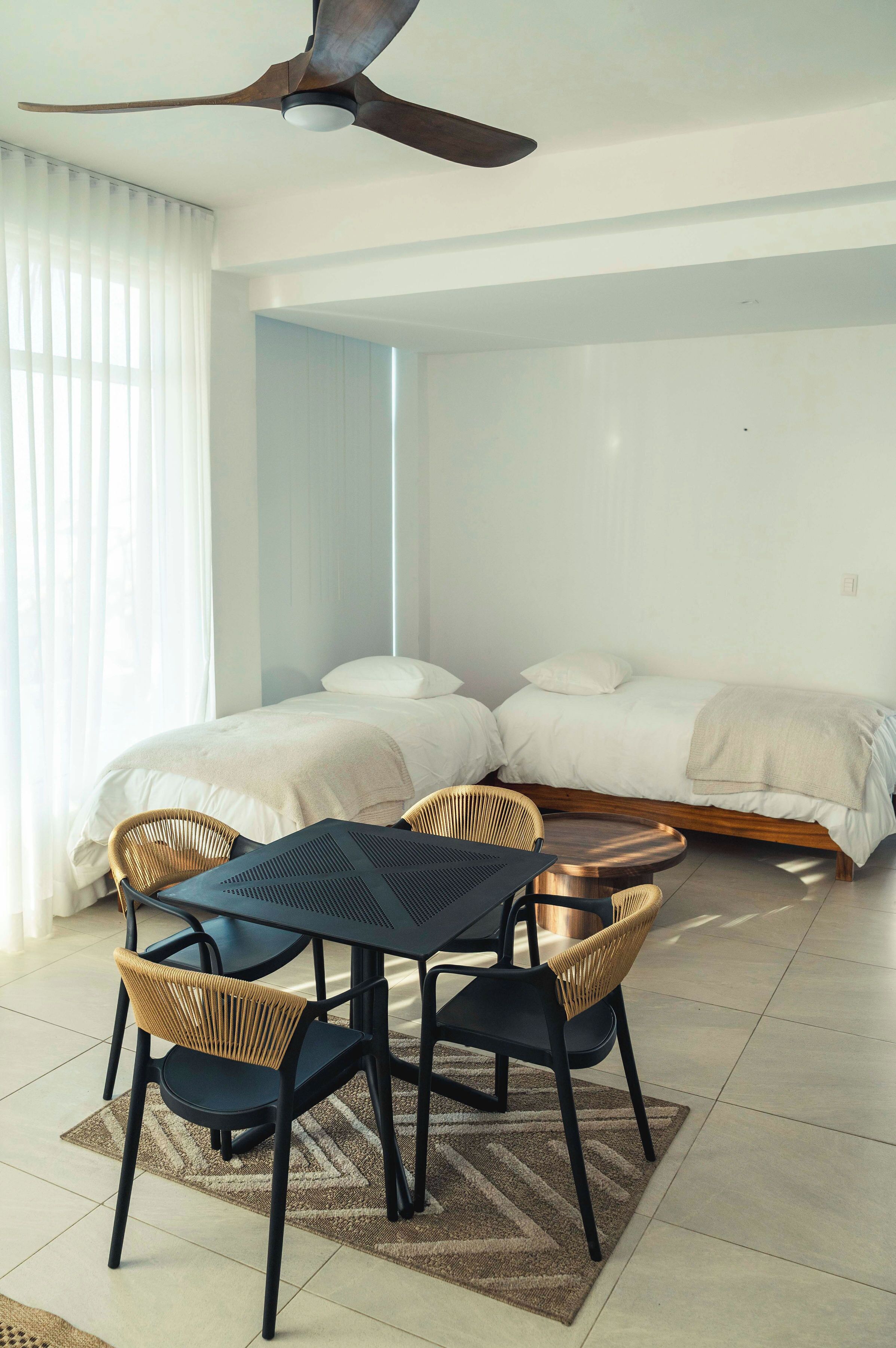 A modern bedroom with two single beds, a ceiling fan, a dining table with chairs, and a large window with sheer curtains.