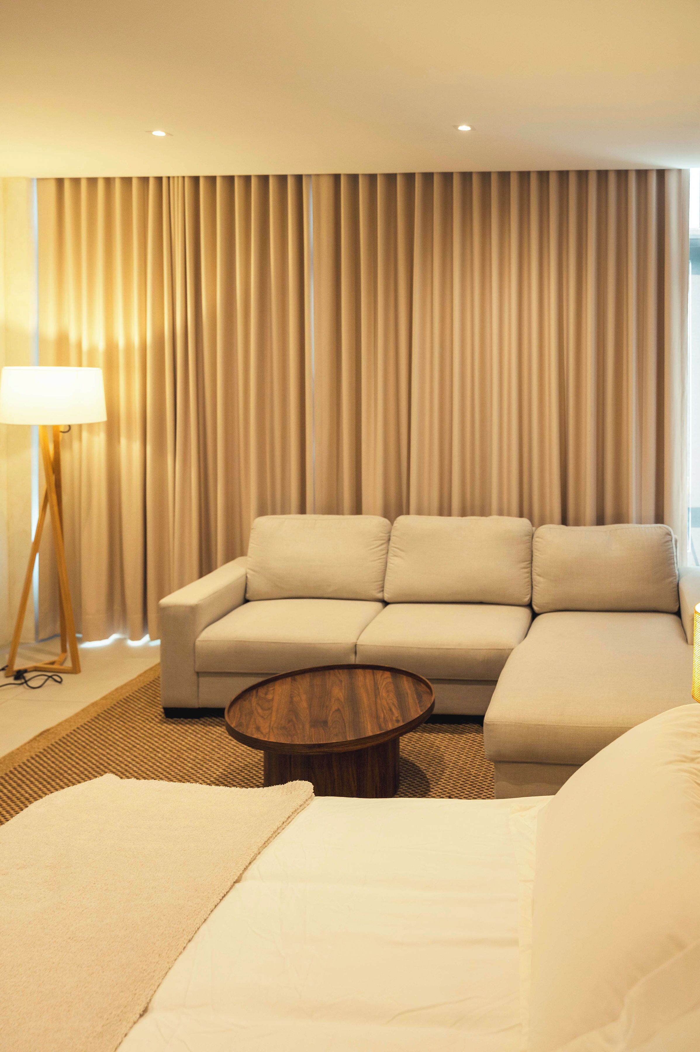 Una habitación de hotel moderna con un sofá color beige, una mesa redonda de madera y un ventanal con cortinas.