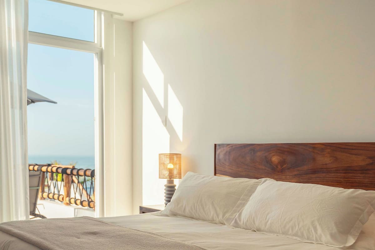 A large bed with white linens, a wooden headboard, and a view of the sea through a large window.