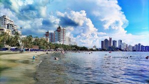 A beach with clear water, palm trees, and buildings in the background. - SGH Hotel Rodadero Suites (Santa Marta)