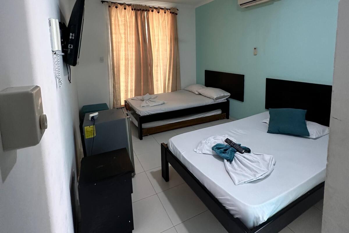 Ein Hotelzimmer mit zwei Betten, einem Deckenventilator, einer Klimaanlage, einem an der Wand montierten Fernseher und einem Fenster mit Vorhängen.