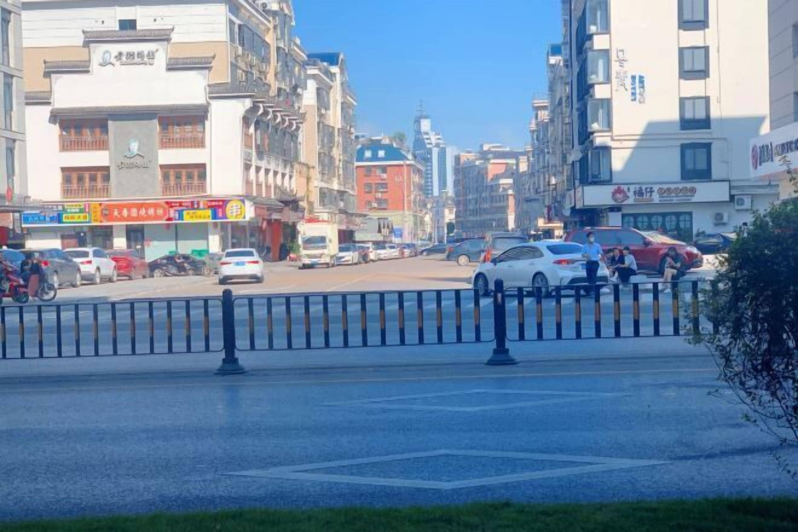 Une rue animée en milieu urbain, bordée de différents bâtiments, dont un bâtiment blanc sur lequel on peut voir une enseigne dans une langue étrangère.