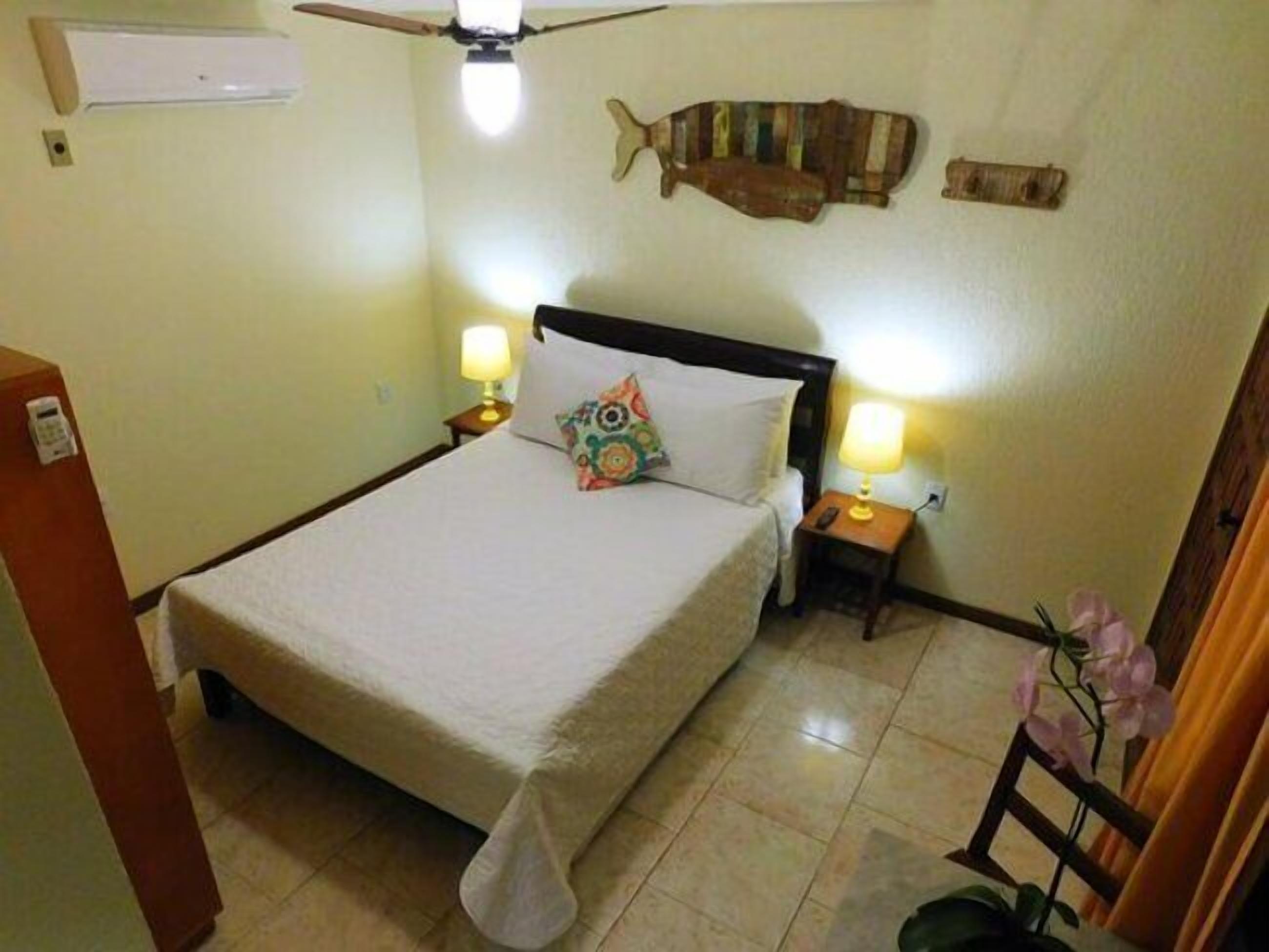 A hotel room with a bed, two bedside lamps, a ceiling fan, a fish-shaped wall decoration, and a small potted plant.