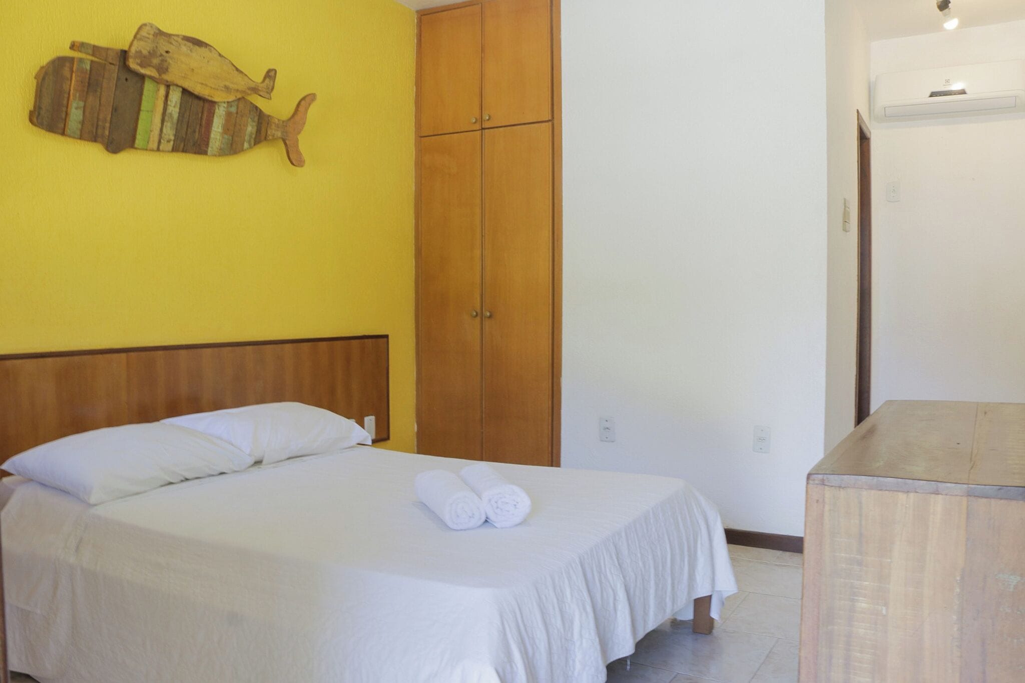 A neatly made bed with white linens and rolled towels, a wooden wardrobe, and a wall-mounted fish decoration.