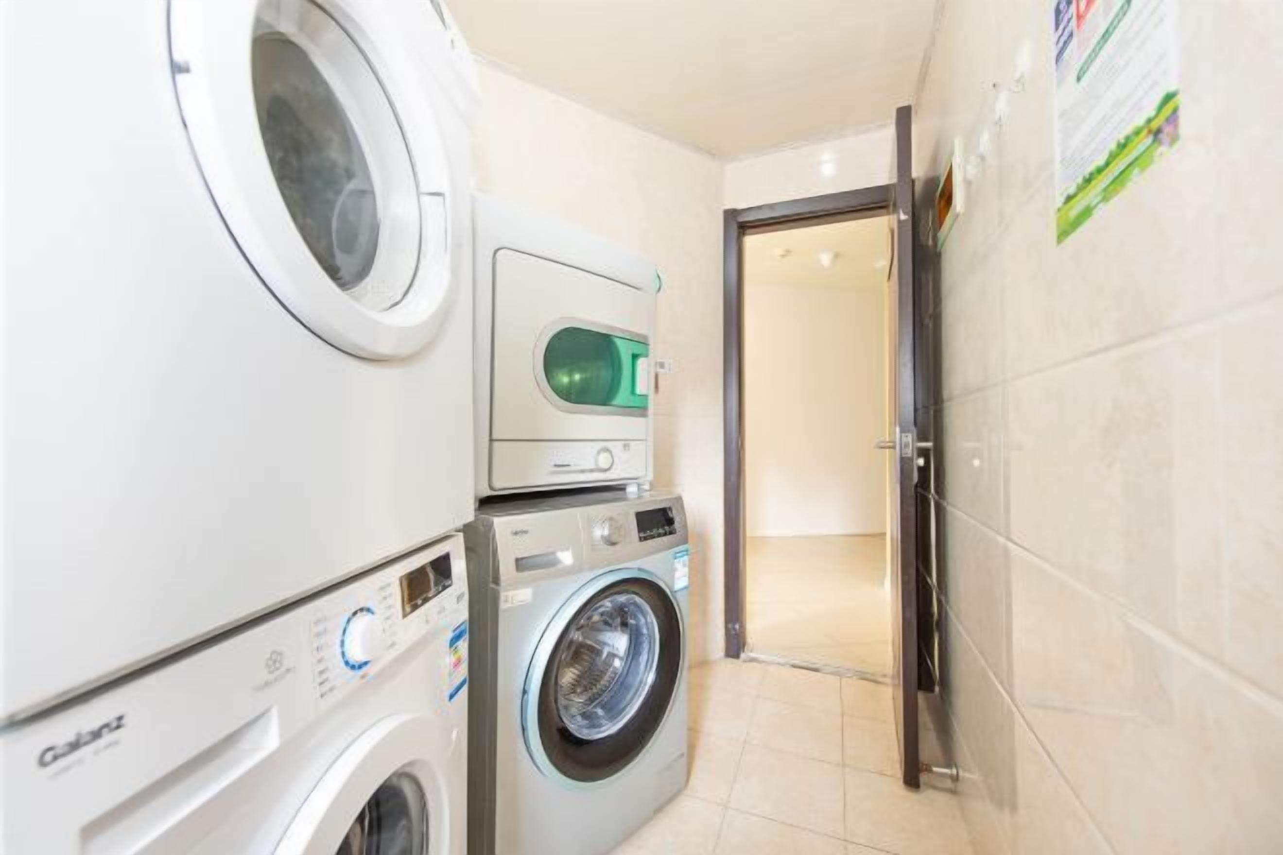 Uma lavanderia com máquina de lavar e secar de carregamento frontal, secadora de parede e uma porta que dá para outro cômodo.