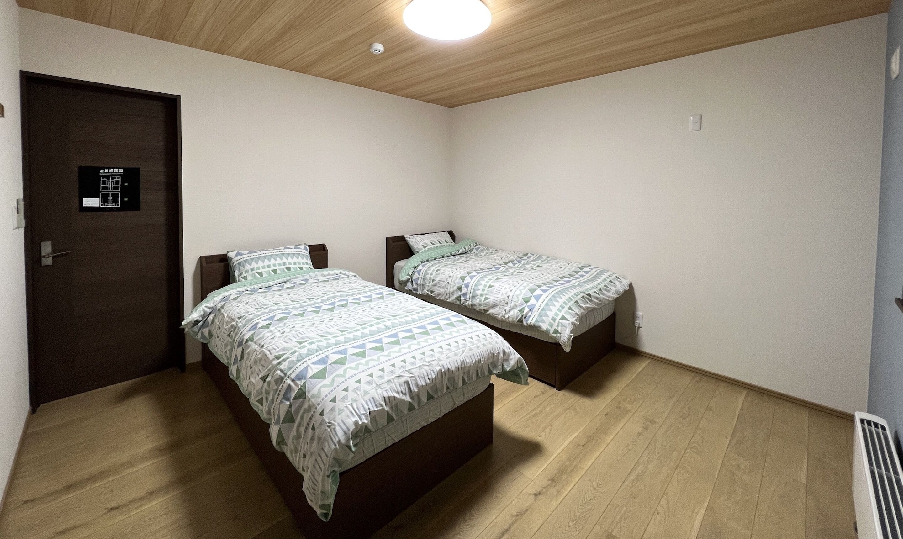 A small, clean room with two beds, a wooden ceiling, and a door with a security panel.